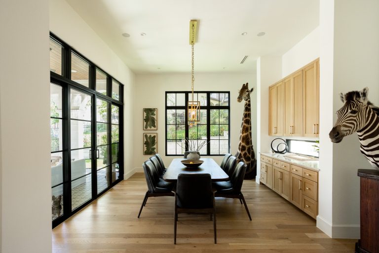 Long image of a dining room with windows, patio door, dining table and chairs, counter, cabinets, and taxidermy zebra and giraffe.